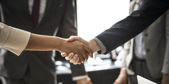Two people shake hands. The handshake is shown in detail, in the background are two people in suits.