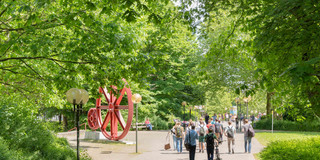 Studierende sind im Sommer auf dem Campus Nord, rote Zahnräder im Hintergrund.